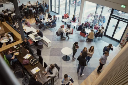 A bustling café with people sitting at tables and standing around, surrounded by large windows.