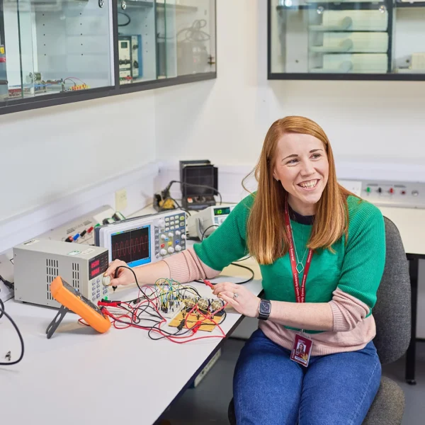 Woman with long red hair sitting at workbench with electronic equipment.