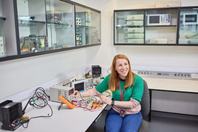 Woman with long red hair sitting at workbench with electronic equipment.