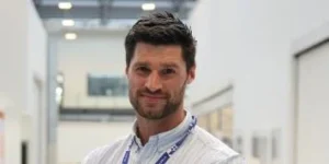 A man with short dark hair and a light blue shirt looks directly at the camera with a slight smile. He is wearing a lanyard around his neck.