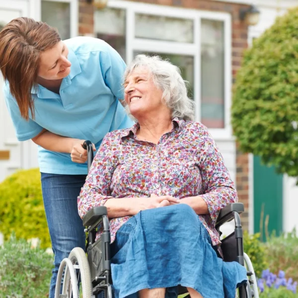 A caregiver assisting an elderly woman in a wheelchair outside a house.