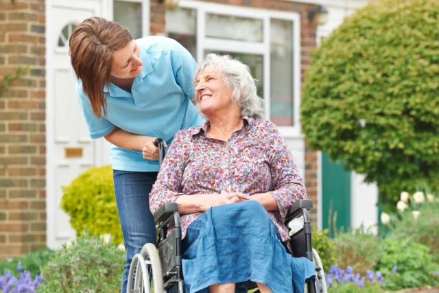 A caregiver assisting an elderly woman in a wheelchair outside a house.