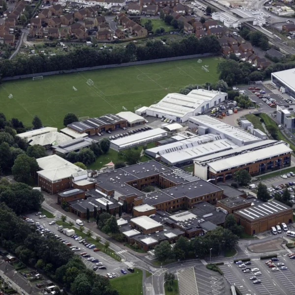 An aerial view of a large complex with several buildings and parking lots, surrounded by trees and a green field.