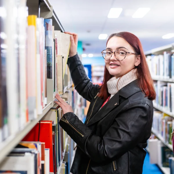 A woman with red hair and glasses browsing through a bookshelf in a library.