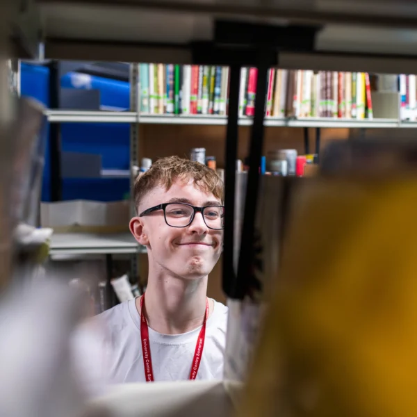 A young man with glasses and a red lanyard looking at books on a shelf.