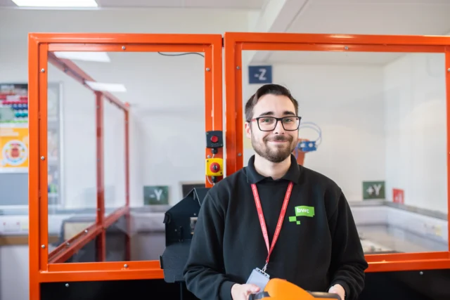 A man with short dark hair, a beard and glasses wearing a black sweatshirt and red lanyard stands in front of glass booths.