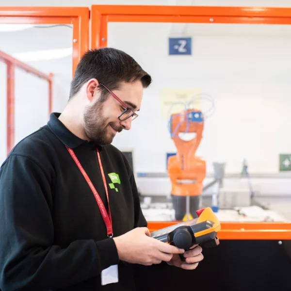 Man in black sweatshirt with red lanyard looking at tablet with robot arm in background.