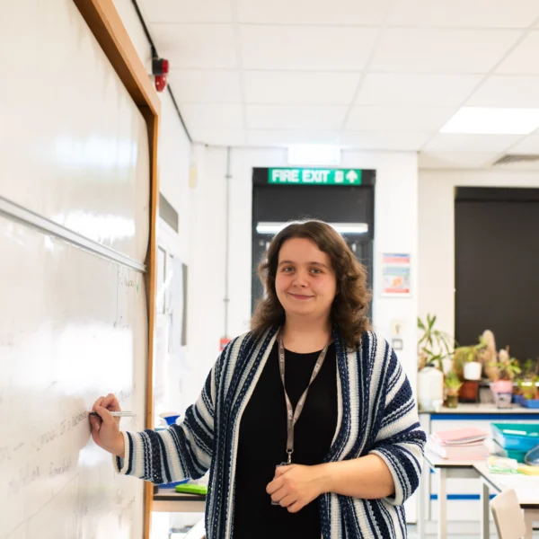 A teacher standing beside a whiteboard in a classroom, smiling at the camera.