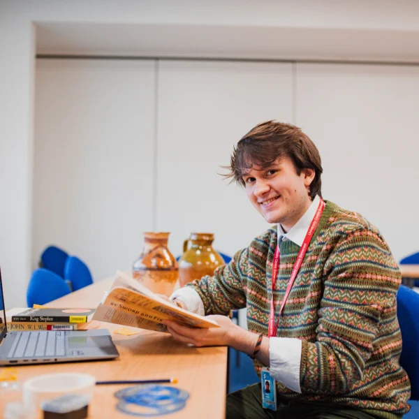 A young person with short brown hair sits at a table with a laptop and book.