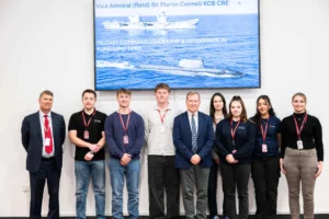 Group of nuclear engineering apprentices and students standing in front of a screen displaying a naval vessel, with Sir Martin Connell.