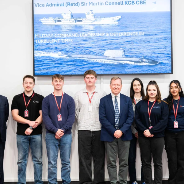 Group of nuclear engineering apprentices and students standing in front of a screen displaying a naval vessel, with Sir Martin Connell.