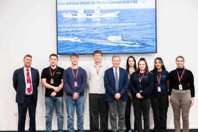Group of nuclear engineering apprentices and students standing in front of a screen displaying a naval vessel, with Sir Martin Connell.