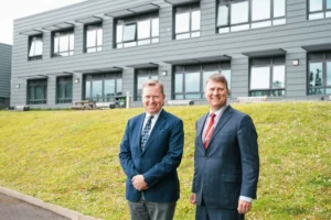Sir Martin Connell and Mike Wenke in suits standing on a grassy hill in front of a modern building which is the National College for Nuclear in Cannington. They are smiling at the camera.