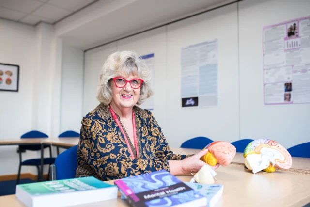 An older woman sitting at a desk with books and a model of the brain.