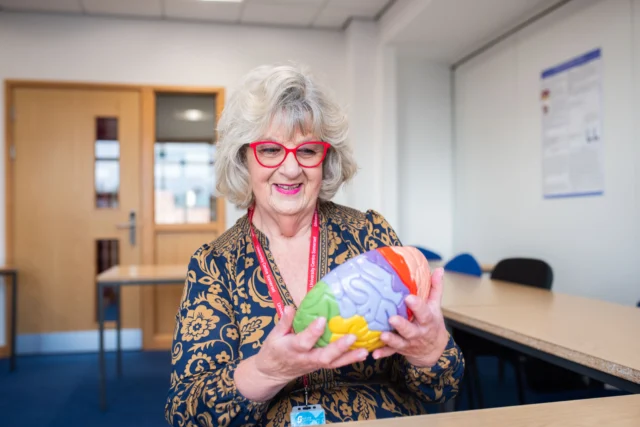 An elderly woman with short gray hair and red glasses holds a puzzle ball. She wears a dark blue top with a gold pattern and a red lanyard.