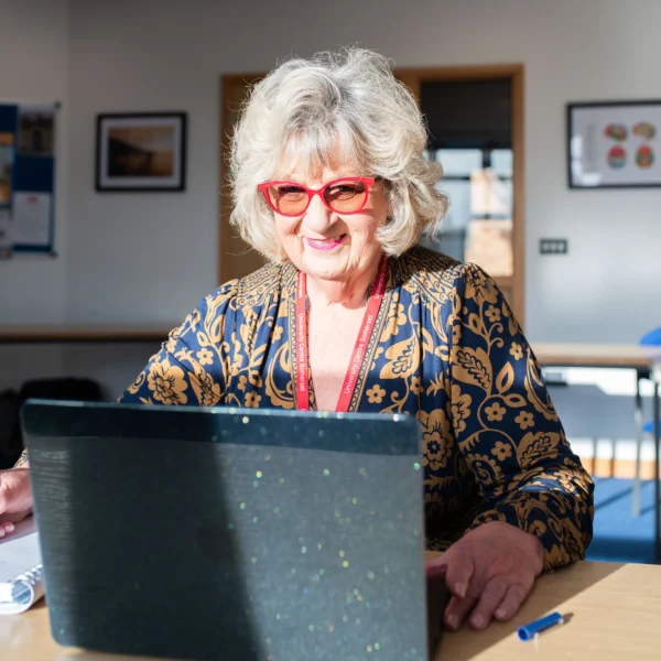 An older woman with short white hair and red glasses sits at a table with a laptop. She appears to be in a classroom or office setting.