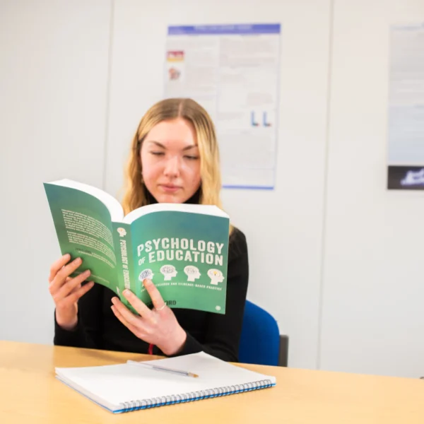 A woman sitting at a desk reading a green book titled 'The Complete CBT Guide'.