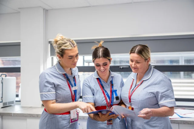 Three female medical staff in light blue uniforms looking at a document.