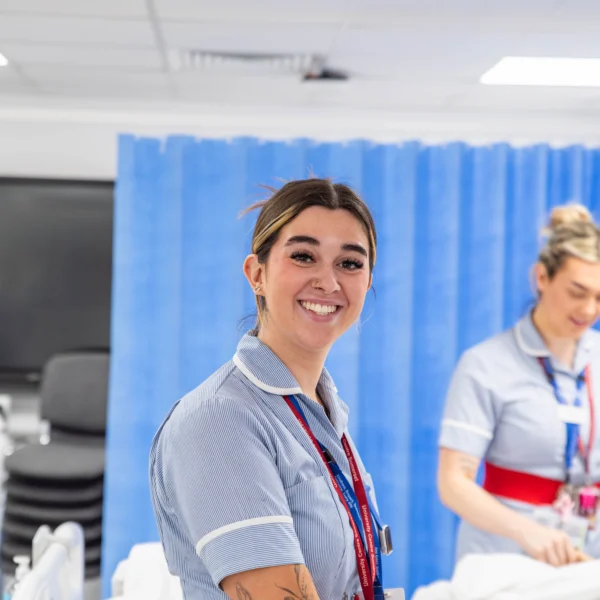 Two female nurses standing in a hospital room, smiling and wearing blue and red uniforms.