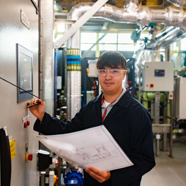 Man in a black coat holding a paper and pressing a button on a control panel in an industrial setting.