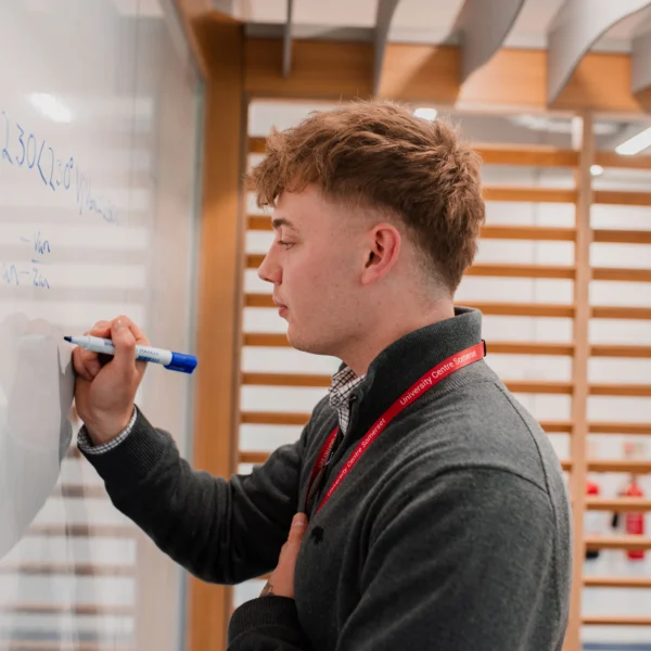 Young man writing on a whiteboard with a blue marker, wearing a gray sweater and red lanyard.