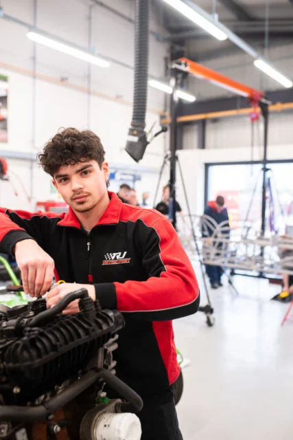A young man in a red and black tracksuit working on an engine.