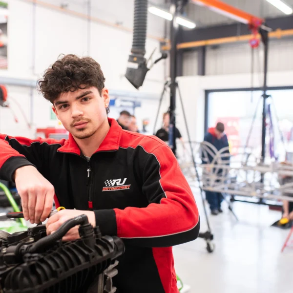 A young man in a red and black tracksuit working on an engine.