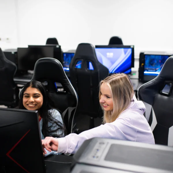 Two young women sitting at computer workstations with racing seats in a classroom setting.