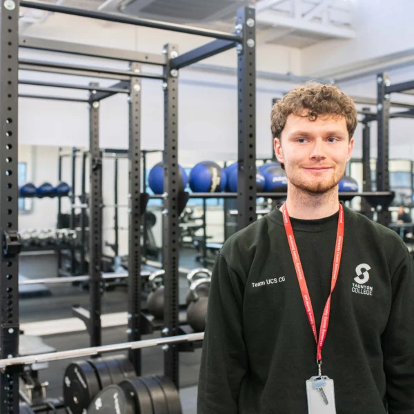 A young man standing in a gym with a red lanyard around his neck, various gym equipment in the background.