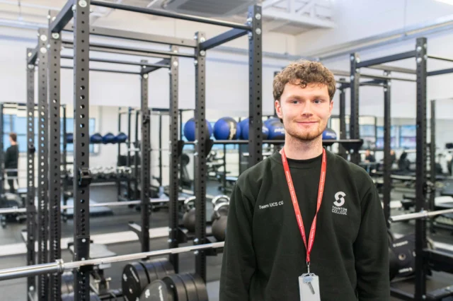 A young man standing in a gym with a red lanyard around his neck, various gym equipment in the background.