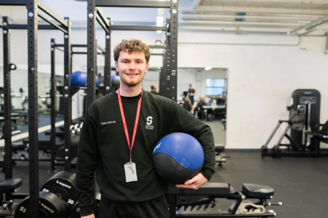 A young man in a gym holding a blue medicine ball.
