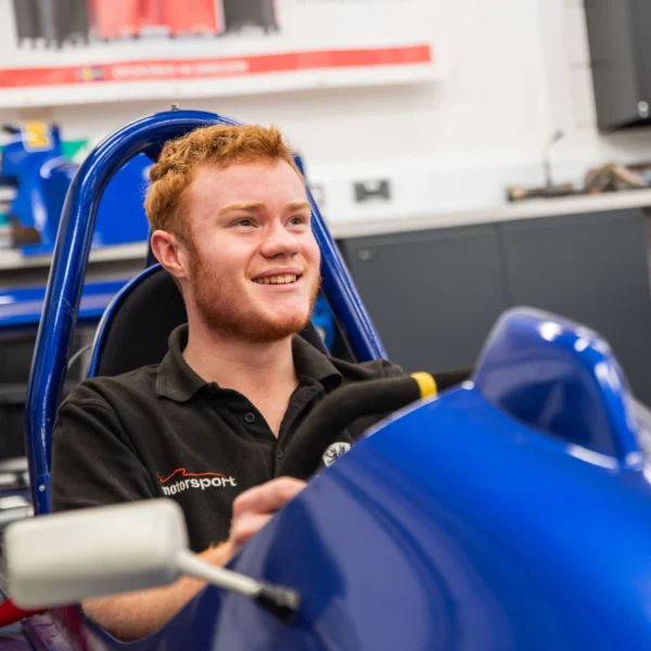 Young man sitting in a blue racing simulator, smiling. He appears to be in a training or testing environment.