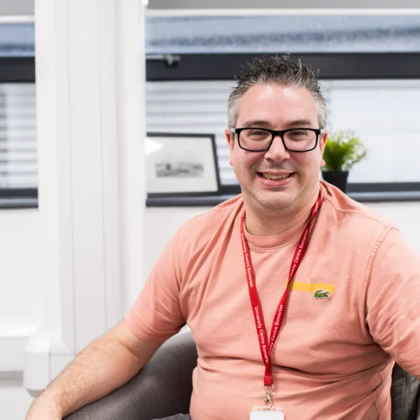 A man sitting in a chair wearing a pink t-shirt and a red lanyard around his neck.