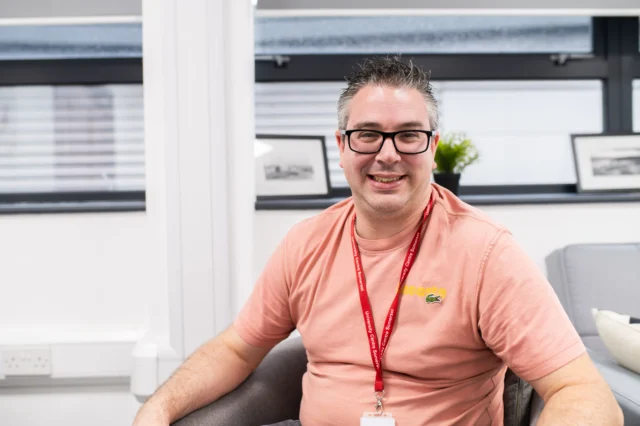 A man sitting in a chair wearing a pink t-shirt and a red lanyard around his neck.