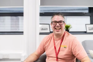 A smiling man wearing a pink shirt and a red lanyard sits in an office.