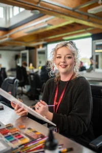 A woman with blonde hair wearing a black sweater and a red lanyard sitting at a desk with art supplies.