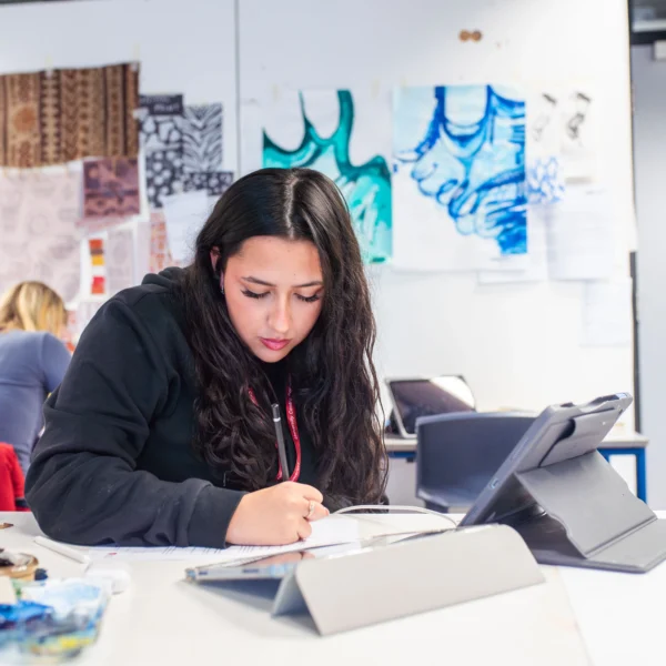 Young woman sitting at a table with a laptop and notebook, surrounded by art supplies and sketches on the wall.