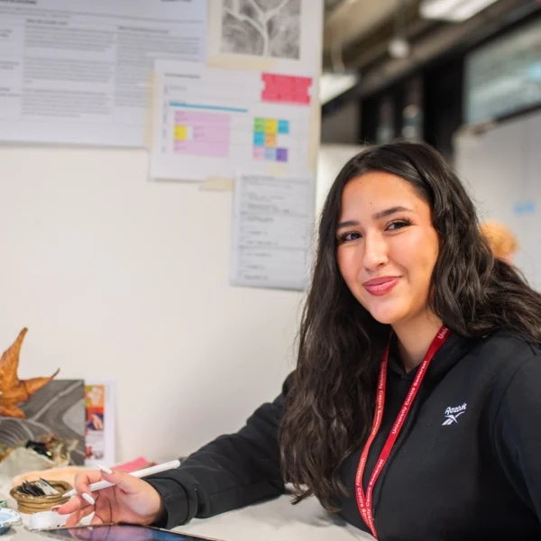 A young woman sitting at a desk with a red lanyard around her neck, surrounded by papers and models.