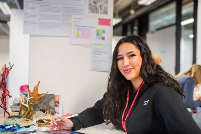 A young woman sitting at a desk with a red lanyard around her neck, surrounded by papers and models.