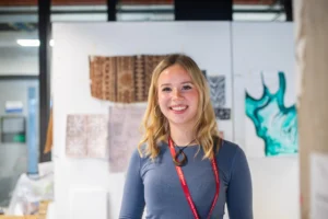A young woman with blonde hair standing in front of artwork on a wall.