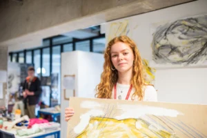 A young woman with long red hair holding a canvas with yellow, white and grey paint.