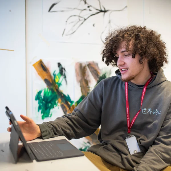 Young man with curly hair sitting at a desk with a laptop and looking at a tablet.