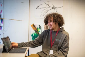 Young man with curly hair using a tablet in a room with a laptop and white wall with paintings.