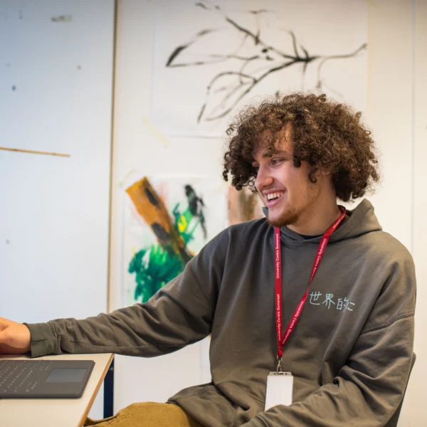 Young man with curly hair using a tablet in a room with a laptop and white wall with paintings.