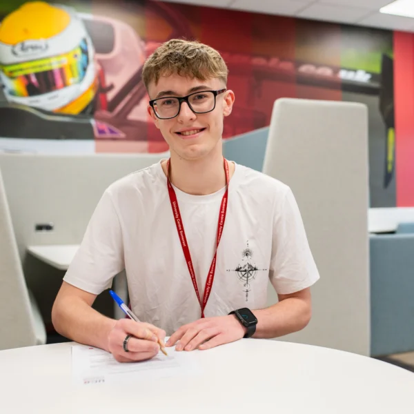 A young man sitting at a table, wearing a white t-shirt and a red lanyard, writing on a piece of paper.