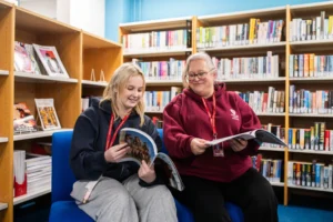 Two women sitting on blue chairs in a library, smiling and reading books.