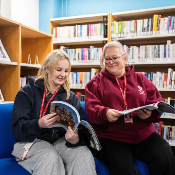 Two women sitting on blue chairs in a library, smiling and reading books.