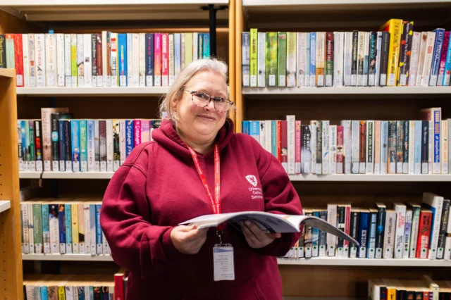 Smiling woman standing in a library, holding an open book.