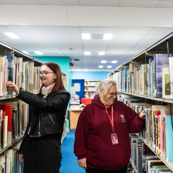 Two women browsing through books on shelves in a library.