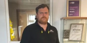 Man in a black polo shirt with a logo on the chest standing in a doorway.
